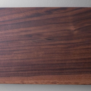 May include: Close-up of a rectangular, dark brown wooden board with visible wood grain. The board has a smooth, polished surface and a natural, warm color. The wood grain pattern creates a textured appearance.