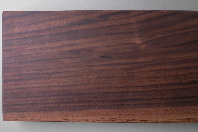 May include: Close-up of a rectangular, dark brown wooden board with visible wood grain. The board has a smooth, polished surface and a natural, warm color. The wood grain pattern creates a textured appearance.