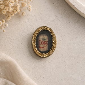 May include: An oval-shaped, gold-colored frame with an ornate border. Inside the frame is a miniature painting of a Diet Coke can. The can is red and white with the words "Diet Coke" visible. The background is black.