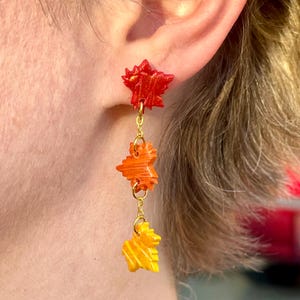 May include: A pair of dangle earrings featuring three layered maple leaf charms. The top charm is red, the middle charm is orange, and the bottom charm is yellow. The charms are connected by a gold chain.