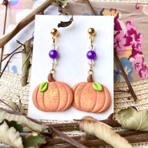 May include: A pair of gold pumpkin earrings with purple and clear beads. The pumpkins are orange with green leaves.