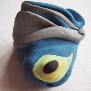 May include: A blue and silver clay sculpture of a bird's head with a yellow and green eye.