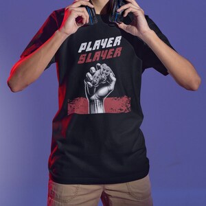 May include: Black t-shirt with the text "PLAYER SLAYER" in white and red. The shirt features a graphic of a fist holding a game controller. The person in the image is wearing headphones.