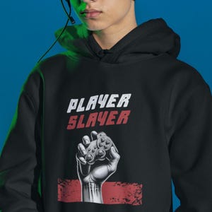 May include: Black hoodie with the words "PLAYER SLAYER" in white and red. The design features a fist holding a video game controller. The hoodie is made of a soft material and has a hood.