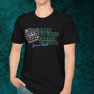 Puede incluir: Camiseta negra con un gráfico de teclado y el texto "BORN TO GAME FORCED TO WORK" en verde y blanco. La camiseta también incluye el texto "Gamers Unite" en una fuente turquesa.