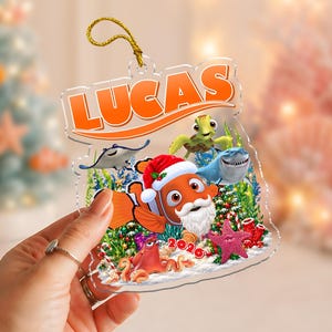 May include: A clear acrylic Christmas ornament featuring the name LUCAS in orange text at the top. The design includes a clownfish wearing a Santa hat and beard, a sea turtle, a shark, a stingray, and an octopus, all set against a festive underwater scene with the year 2026 printed in red.