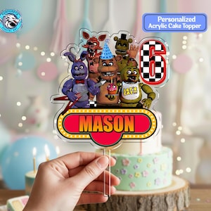 May include: A clear acrylic cake topper featuring cartoon characters, the number 6, and the name "MASON" in a red and yellow design. The topper is held in front of a cake with a wooden base. The text "Personalized Acrylic Cake Topper" is also visible.