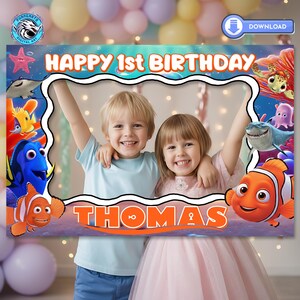 May include: A photo frame with the text "HAPPY 1st BIRTHDAY" and the name "THOMAS". The frame features cartoon characters from the movie "Finding Nemo" and is designed for a birthday celebration. Two children are posing inside the frame.