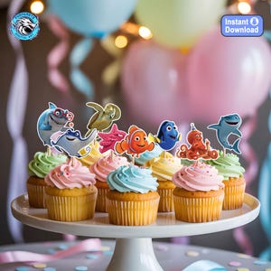 May include: A white cake stand holds cupcakes with colorful frosting. Each cupcake is topped with a character from the movie "Finding Nemo". The background includes balloons in shades of blue, pink, and yellow, with a blurred string of lights.