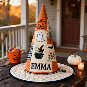 May include: A quilted witch hat featuring orange and cream panels with embroidered Halloween motifs including a jack-o'-lantern, ghost, spider web, bat, and a cat in a cauldron. The name EMMA is embroidered in black on the front base panel.