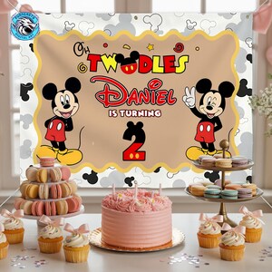 May include: A rectangular birthday backdrop featuring Mickey Mouse, the text Oh Twodles, Daniel Is Turning 2, and a scalloped tan center. The banner is displayed behind a pink frosted cake, a macaron tower, and a tiered tray of colorful macarons.