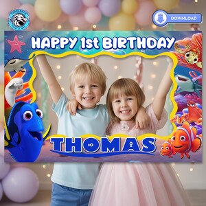 May include: A photo booth frame featuring the text "HAPPY 1st BIRTHDAY" and the name "THOMAS". The frame is decorated with cartoon characters from the movie "Finding Nemo" and "Finding Dory". Two children are posing inside the frame.
