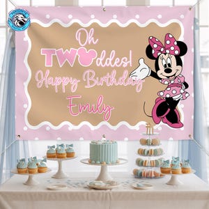 May include: A rectangular pink and tan birthday banner featuring a cartoon mouse in a polka dot dress, the text Oh TWOddes! Happy Birthday Emily, and metal grommets in the corners. The banner is displayed above a table with a cake and desserts.