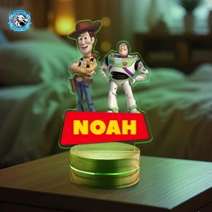 May include: A night light featuring Woody and Buzz Lightyear figures. The figures stand on a red base with the name "NOAH" in yellow. The base is illuminated with a green glow, and the light sits on a wooden surface.