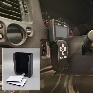May include: A black plastic phone holder with adhesive strips for mounting. The holder is designed to fit a small electronic device, such as a phone or a GPS unit. The holder is mounted on the dashboard of a car.