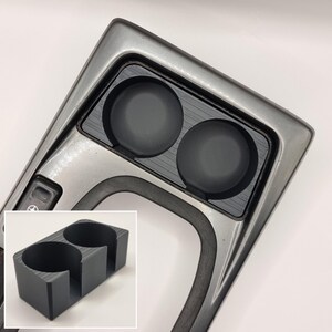 May include: A black plastic cup holder with two circular openings. The cup holder is designed to fit into a car's center console.