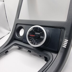 May include: A black plastic car dashboard with a silver TurboSmart 2.0 boost gauge. The gauge has a black face with white markings and a red needle.
