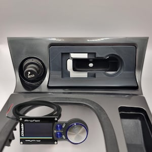 May include: A black and gray Pro-Fec aftermarket electronic boost controller with a digital display and a knob with a blue ring. The controller is mounted in a gray car interior with a cigarette lighter and a phone holder.
