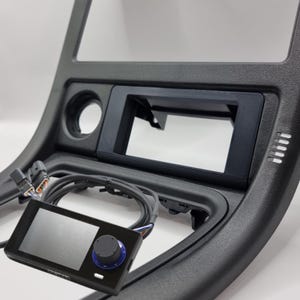 Könnte beinhalten: Ein schwarzes Armaturenbrett-Kit für Autos mit einem leeren Bildschirm und einem schwarzen Bedienfeld mit einem blauen Knopf. Das Kit enthält einen Kabelbaum.