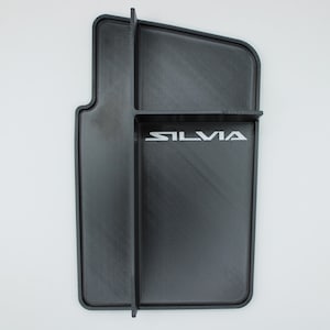 May include: A black car console organizer with the word "SILVIA" in white lettering. The organizer has a rectangular shape with a curved edge and a built-in divider. The product is designed to fit in a car.