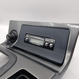 May include: A black car dashboard with a cigarette lighter and a digital clock radio with the text "Grectio Full Auto Timer" on the display.