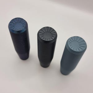 Könnte beinhalten: Drei Schaltknäufe in verschiedenen Farben. Die Knäufe sind schwarz, blau und grau. Jeder Knauf hat eine strukturierte Oberfläche und ein kreisförmiges Design auf der Oberseite.