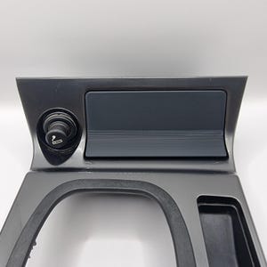 May include: Black plastic car interior trim piece with a cigarette lighter, a small storage compartment, and a larger storage compartment.