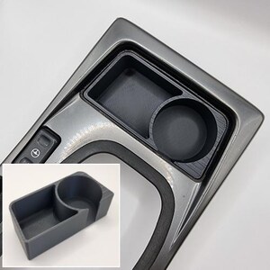 May include: A gray plastic cup holder with two compartments. The cup holder is designed to fit into a car's center console.