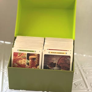 May include: A green recipe box with a lime green lid. The box is open and filled with recipe cards. The cards have pictures of food on them. The top card on the left side reads 