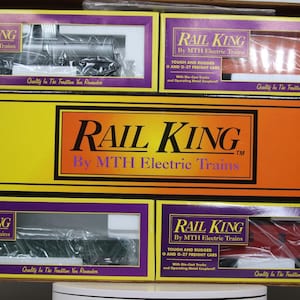 Puede incluir: Se muestran varias cajas de trenes eléctricos Rail King. Las cajas son amarillas, naranjas y moradas, con el logotipo de Rail King. Las cajas contienen modelos de vagones de tren, incluyendo una locomotora y vagones de carga.