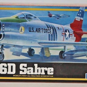 Puede incluir: Caja de maqueta del F-86D Sabre, un interceptor para todo clima de la Fuerza Aérea de EE. UU. La ilustración muestra un avión plateado y rojo con el texto "U.S. AIR FORCE". El kit es a escala 1/72 y es adecuado para niños de 10 años en adelante.