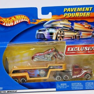May include: Hot Wheels Pavement Pounder toy set. The set includes a red motorcycle, a yellow and black trailer with a flame design, and a red truck. The packaging is blue with the Hot Wheels logo and the word "EXCLUSIVE".