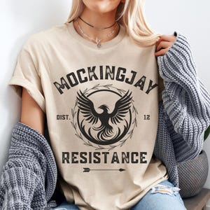 May include: A beige t-shirt with a black graphic print of a phoenix with the text "MOCKINGJAY DIST. 12 RESISTANCE" and an arrow.