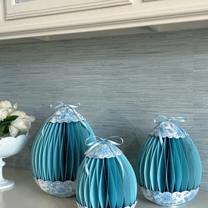 Puede incluir: Tres adornos decorativos de papel con forma de huevo de diferentes tamaños. Los huevos son de color azul claro con un diseño plisado y un estampado floral blanco en la parte superior e inferior. Cada huevo está rematado con un lazo de cinta azul claro.