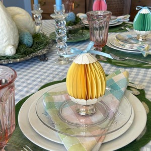 May include: A festive Easter table setting with a yellow paper egg decoration on a silver stand. The table is set with white plates, clear glassware, and a pastel-colored checkered napkin. Blue candles and a bunny figurine are also visible.