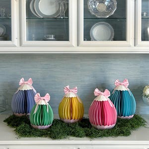 Puede incluir: Cinco decoraciones de huevos de papel de colores con lazos rosas, dispuestas sobre musgo verde. Los huevos son en tonos de azul, verde, amarillo, rosa y turquesa. El fondo presenta armarios blancos y una pared azul claro.
