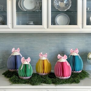 May include: Five colorful paper egg decorations with pink bows on top, arranged on a bed of green moss. The eggs are in shades of blue, green, yellow, pink, and teal. The background features white cabinets and a light blue wall.