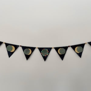 Puede incluir: Una pancarta negra y dorada con un diseño de luna y sol. La pancarta está hecha de tela y tiene una cuerda de cuentas de madera que la atraviesa.