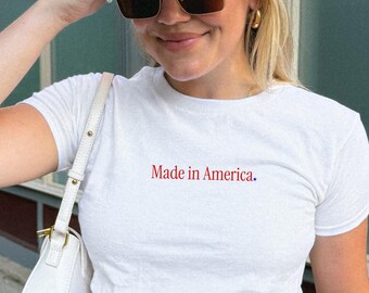 May include: A white short-sleeved t-shirt with the text Made in America. printed in red on the chest, paired with a white baseball cap featuring dark text, dark-tinted sunglasses, and a white shoulder bag with gold-tone hardware accents.