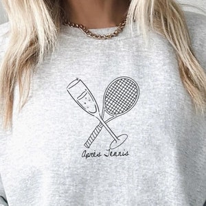 Puede incluir: Una sudadera gris claro con un gráfico negro de una copa de champán y una raqueta de tenis cruzadas. Las palabras "Après Tennis" están impresas debajo. La sudadera es de estilo casual.