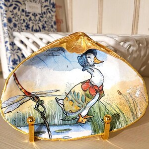May include: A decorative seashell dish with a gold rim, featuring a painted illustration of a duck wearing a blue bonnet and a red bow, and a dragonfly. The dish is on a gold stand.