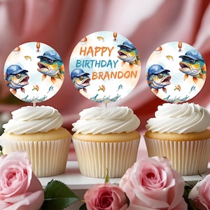 May include: Three cupcakes with white frosting and decorative toppers. The toppers feature a watercolor illustration of a fish wearing a blue cap and sunglasses, with the text "HAPPY BIRTHDAY BRANDON". Pink roses are in the foreground.