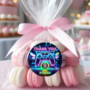 May include: A cellophane bag tied with a pink ribbon, filled with pink and white macarons. A round sticker on the bag reads "THANK YOU" with a neon video game controller graphic and the name "ATHENA".