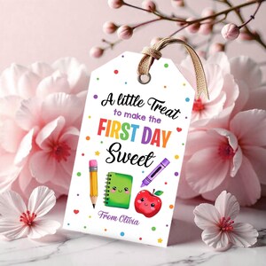 Puede incluir: Una etiqueta de regalo rectangular blanca con un cordel beige, decorada con puntos de colores y el texto A little Treat to make the FIRST DAY Sweet. Las ilustraciones incluyen un lápiz amarillo, un cuaderno verde con cara, un crayón morado y una manzana roja.