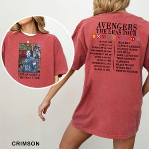 May include: Crimson-colored t-shirt featuring a Captain America graphic on the front and "Captain America The Eras Tour" text. The back displays "Avengers The Eras Tour" with character names and dates.