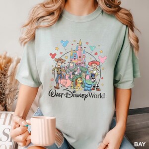 May include: A light green t-shirt featuring a colorful graphic of Disney's Toy Story characters, including Woody, Buzz Lightyear, and Jessie, in front of a castle. The words "Walt Disney World" are printed below the characters.