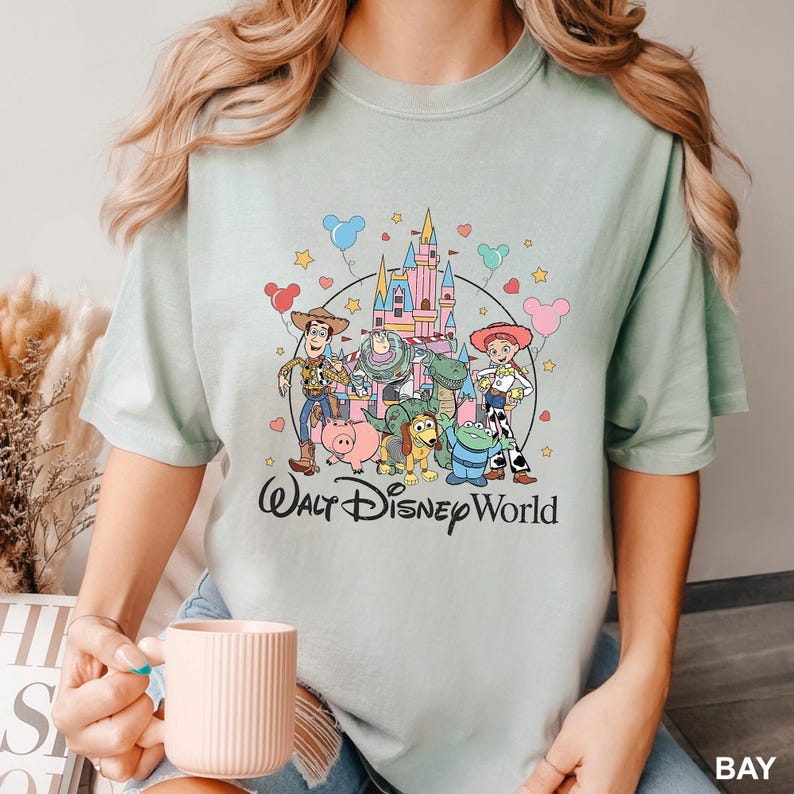 May include: A light green t-shirt featuring a colorful graphic of Disney's Toy Story characters, including Woody, Buzz Lightyear, and Jessie, in front of a castle. The words "Walt Disney World" are printed below the characters.