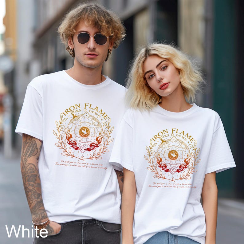 May include: A white short-sleeved t-shirt featuring a circular gold and red graphic with the text Iron Flame Turn It Down. The design includes a central bird emblem, surrounding laurel leaves, and flames, with small text printed below the graphic.
