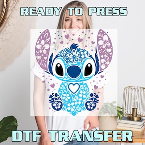May include: A white t-shirt with a blue and purple floral Stitch design. The design features a blue Stitch with a heart on its chest and a floral pattern around it. The text "READY TO PRESS" is at the top of the image and "DTF TRANSFER" is at the bottom.