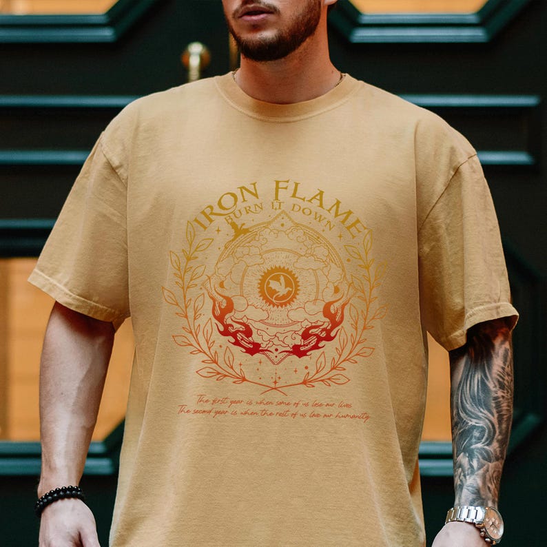 May include: A tan short-sleeved t-shirt featuring a circular graphic with the text Iron Flame Burn It Down. The design includes a central dragon emblem, flames, and laurel branches in orange and red, with small text printed at the bottom.